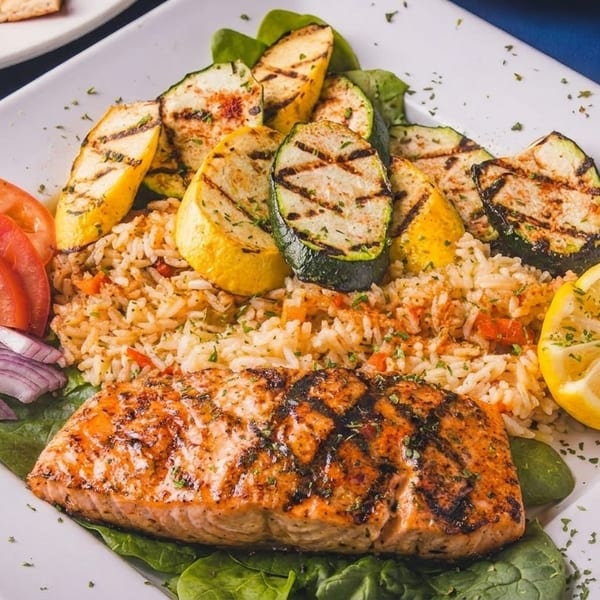 Grilled salmon with rice, zucchini, squash, tomatoes, red onions, and a lemon wedge.